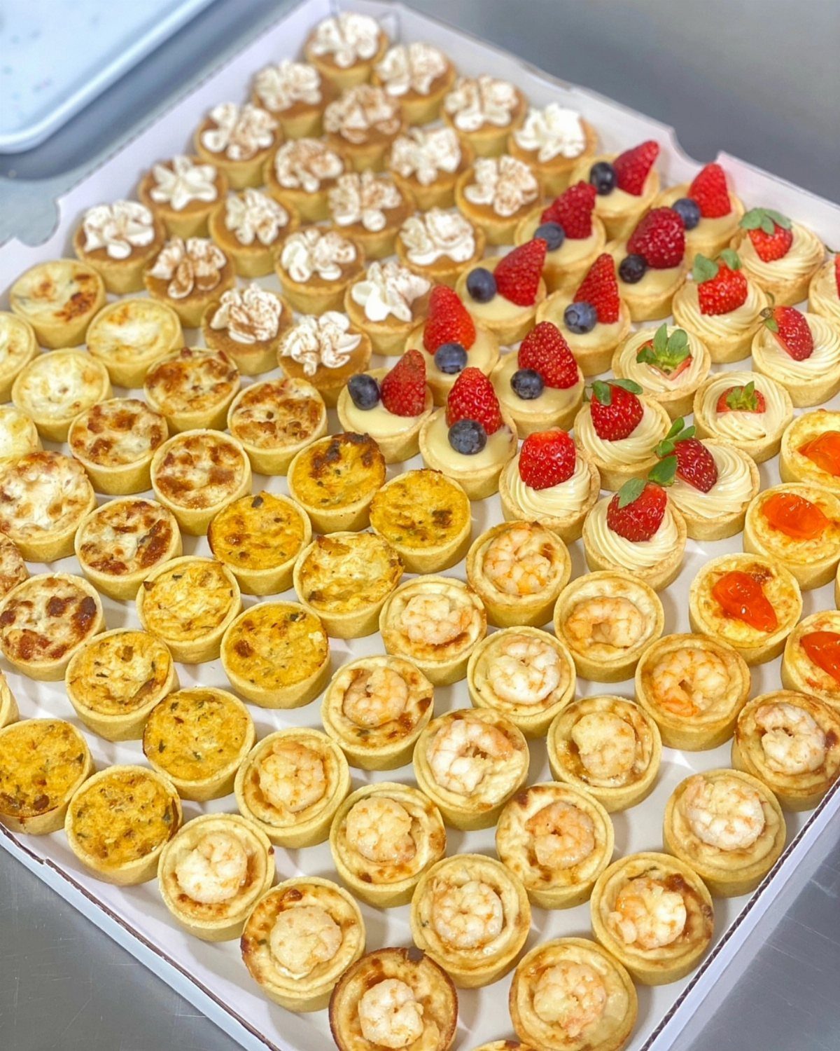 Miniature savory quiches and fruit pastries arranged in a white catering box.