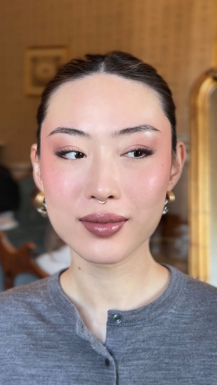 Woman with radiant glowy skin, pink eyeshadow, and glossy mauve lips posing.