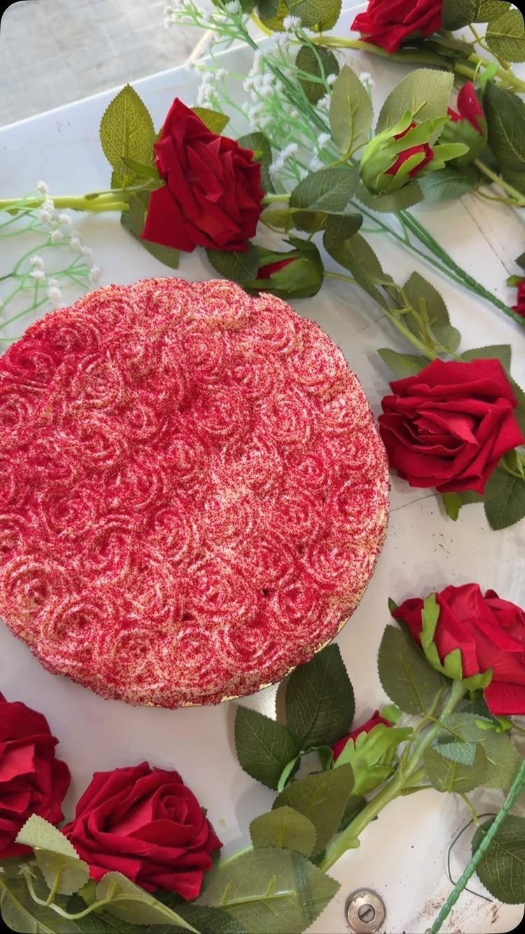 Red rosette frosted cake surrounded by red roses and green leaves on white.