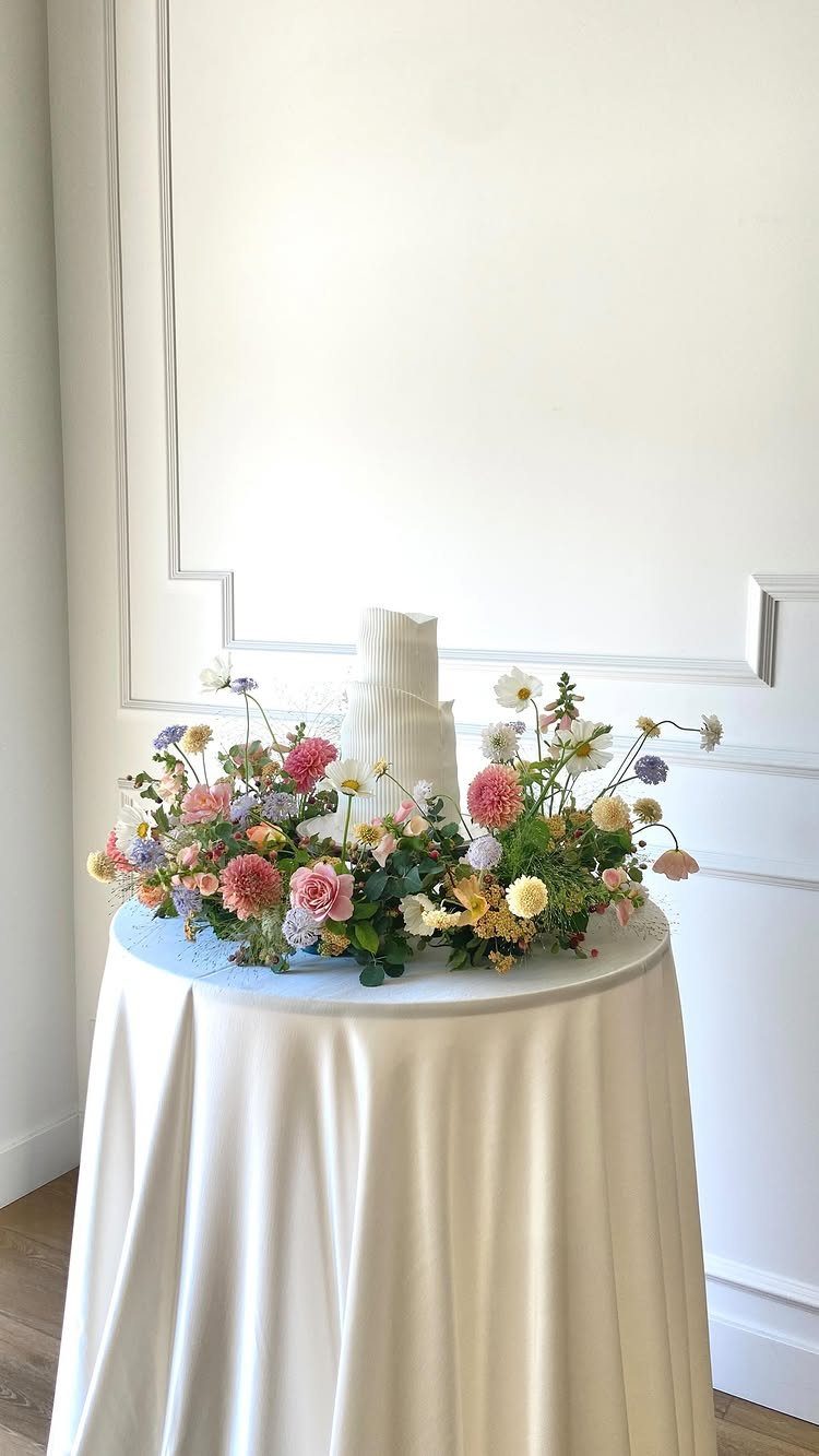 White tiered wedding cake surrounded by colorful fresh flowers on a white table.