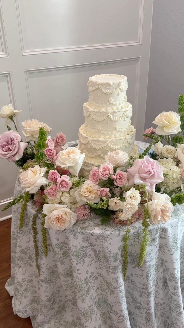 Three-tier white wedding cake surrounded by pink and cream roses on a patterned tablecloth