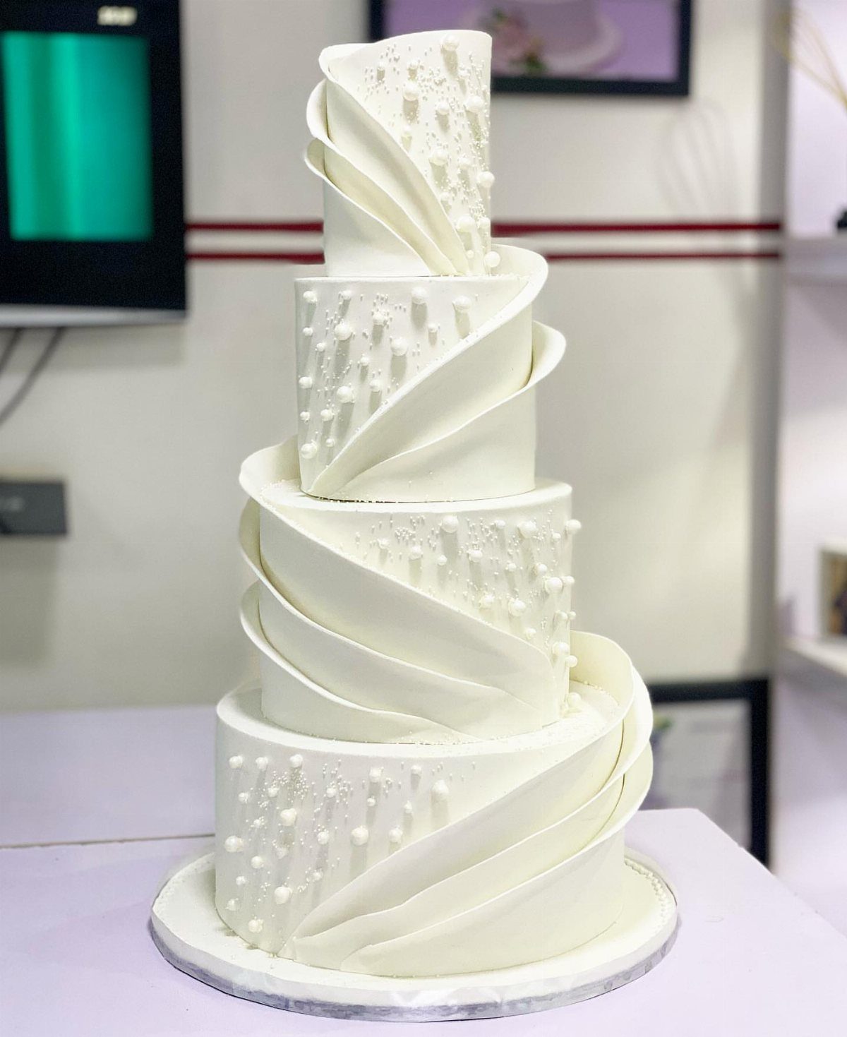 Four-tier white wedding cake with swirling fondant ribbons and pearl decorations.