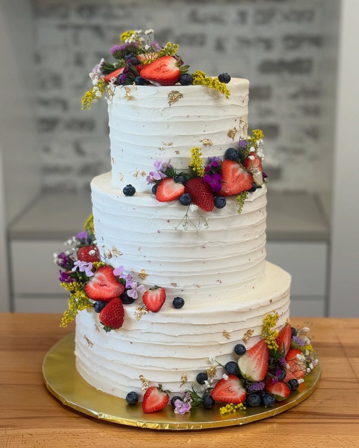 Three-tier white wedding cake decorated with fresh strawberries, blueberries, and colorful wildflowers.