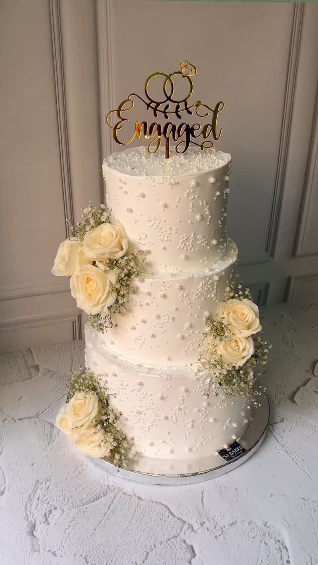 Three-tier white cake with cream roses and a gold Engaged topper.