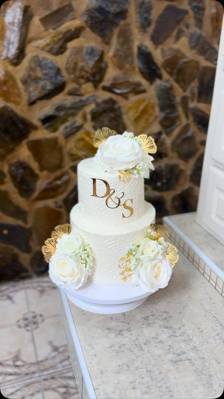 Two-tier white wedding cake with gold initials and white roses on a stand.