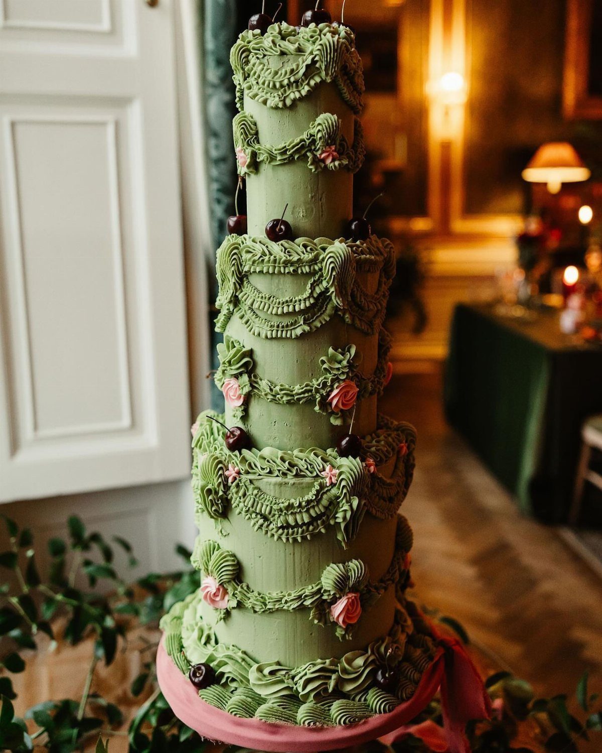 Multi-tiered green wedding cake with ornate frosting, pink flowers, and dark cherries.