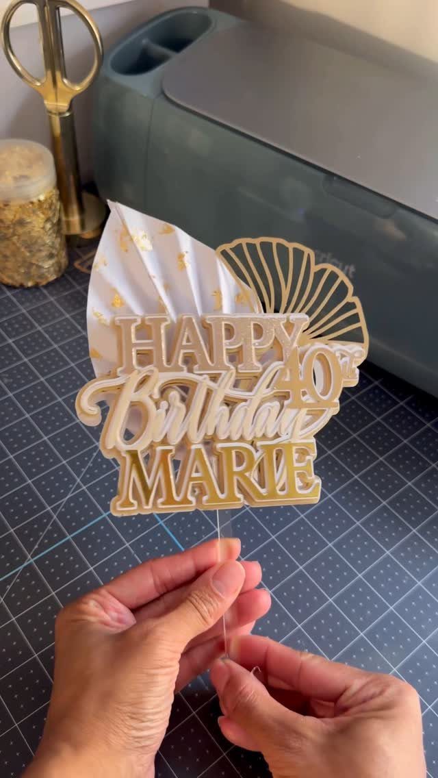 Gold and white personalized cake topper reading Happy 40th Birthday Marie held by hands.