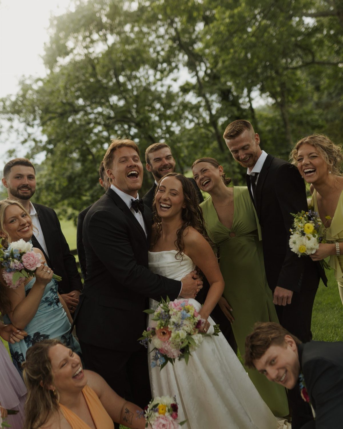 Laughing wedding party with bride and groom embraced outdoors among green trees.