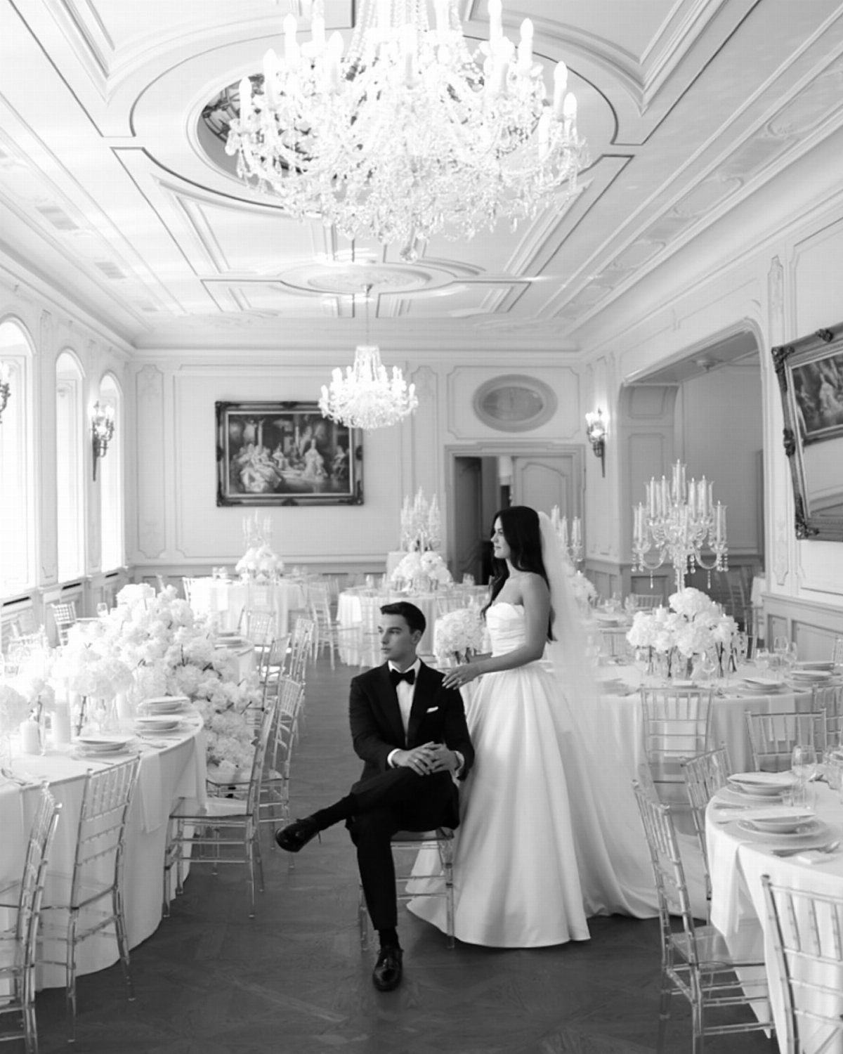 Bride and groom posing in an elegant ballroom with crystal chandeliers and white flowers.