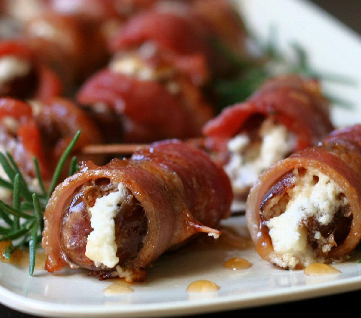Bacon-wrapped dates stuffed with goat cheese on a white plate with rosemary.