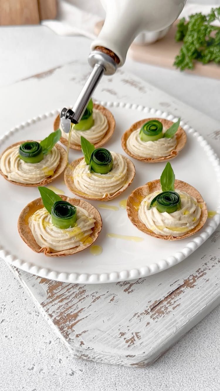 Hummus and veggie cups with cucumber rolls and basil on a white plate.