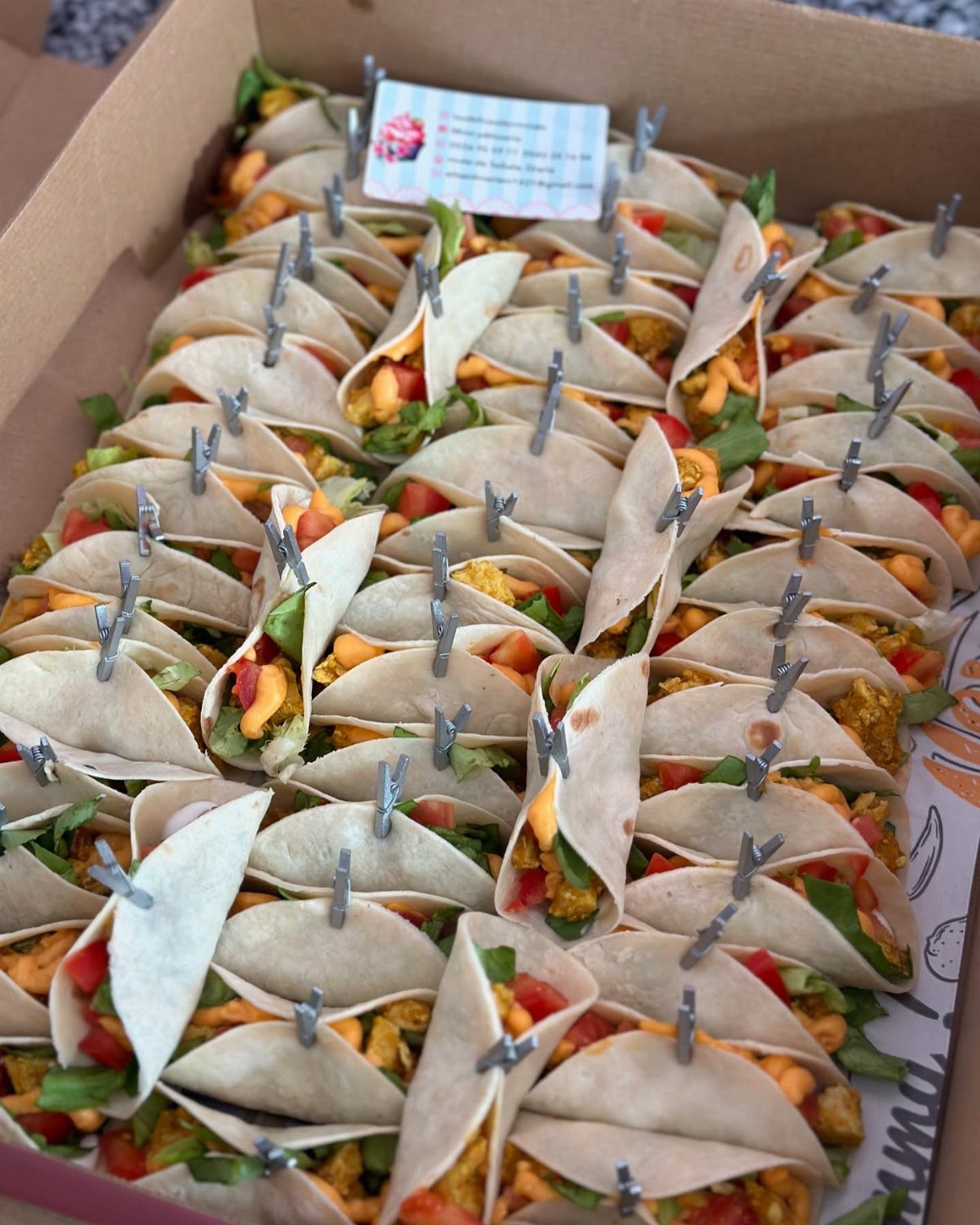 Mini tacos with colorful fillings held open by silver clips in a box.
