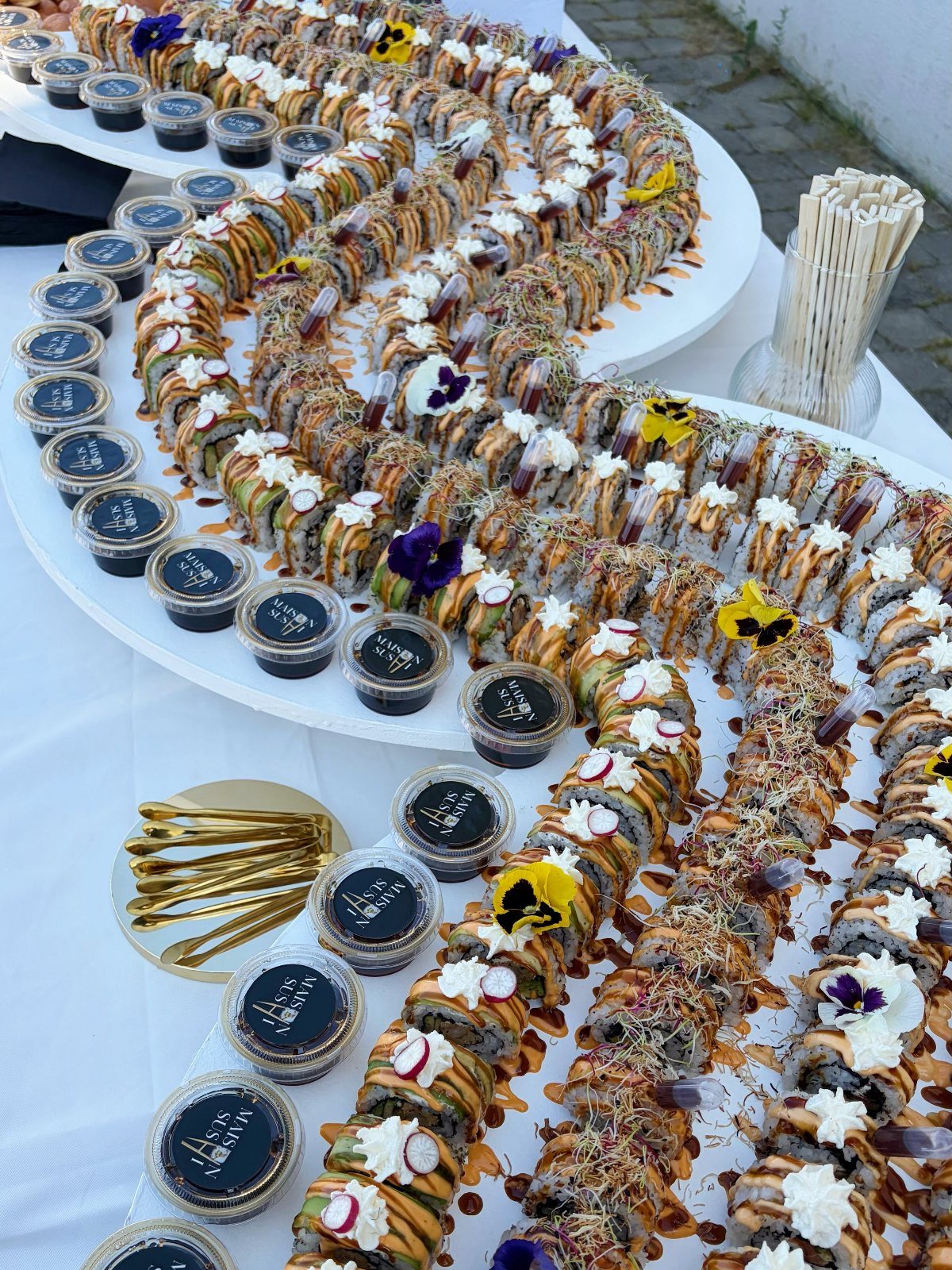 Sushi grazing table with assorted rolls, dipping sauces, and gold forks on white.