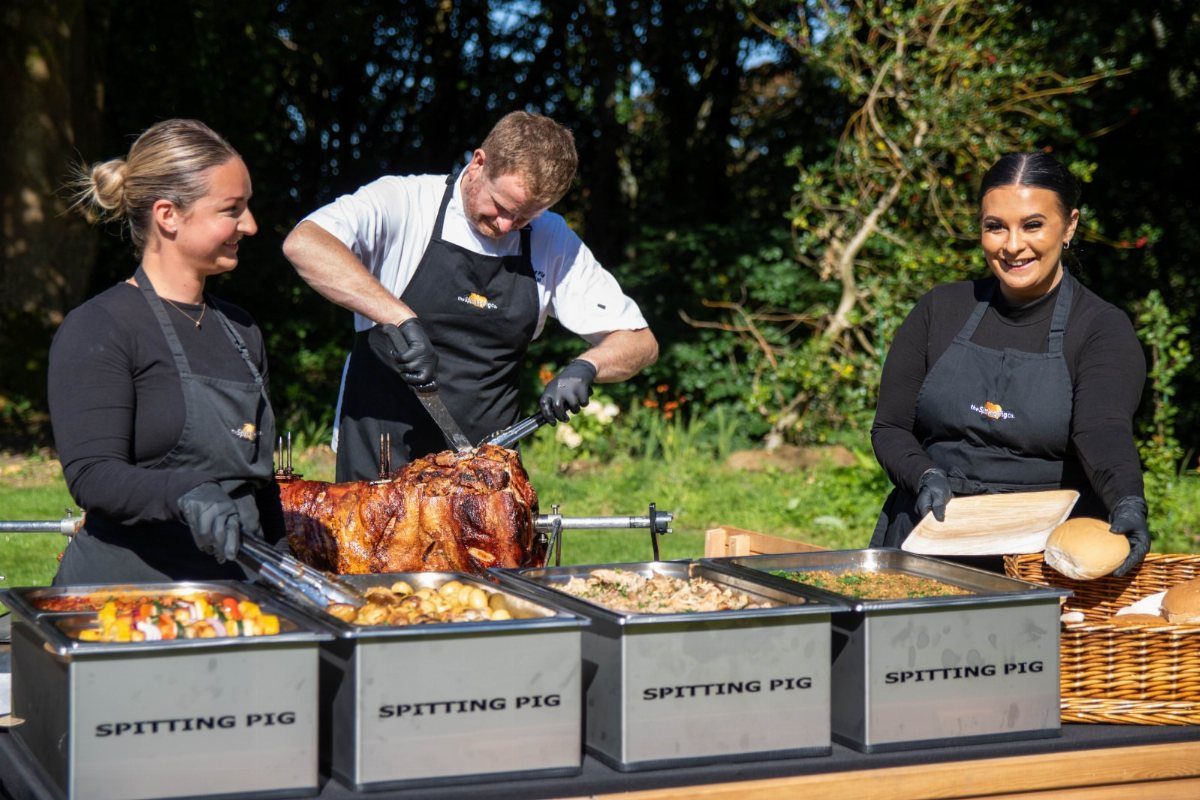 Catering staff carving a whole roasted pig on a spit with buffet side dishes.