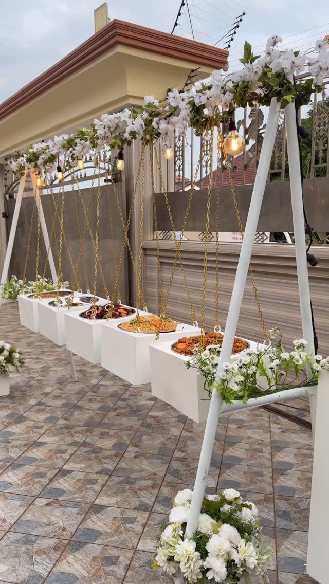 White pedestals with food platters under a floral frame with hanging lights.