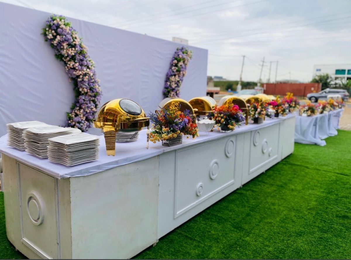 White buffet table with gold chafing dishes and flowers on green grass