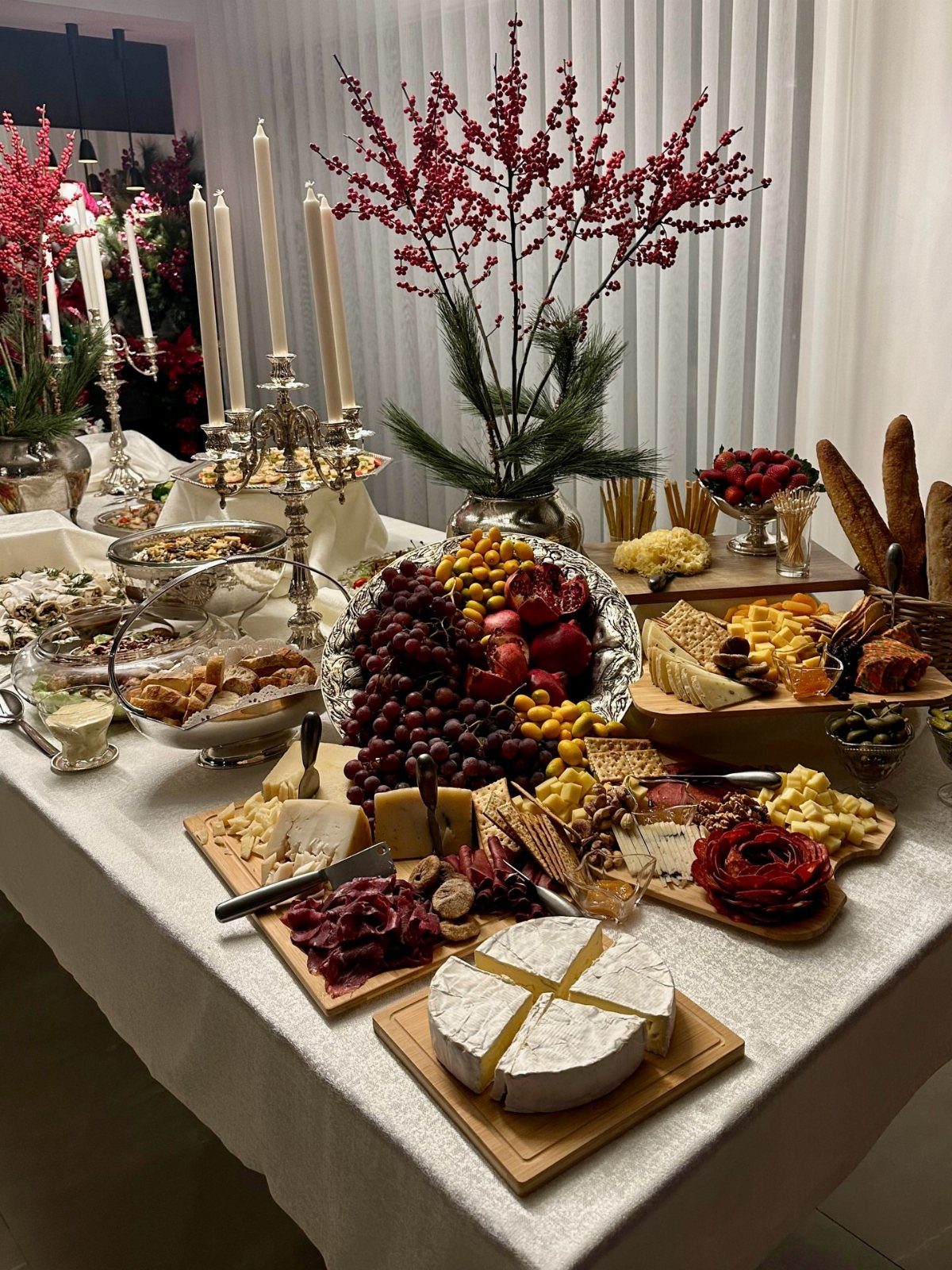 Buffet table with cheese and fruit platters decorated with white candles and red berries.