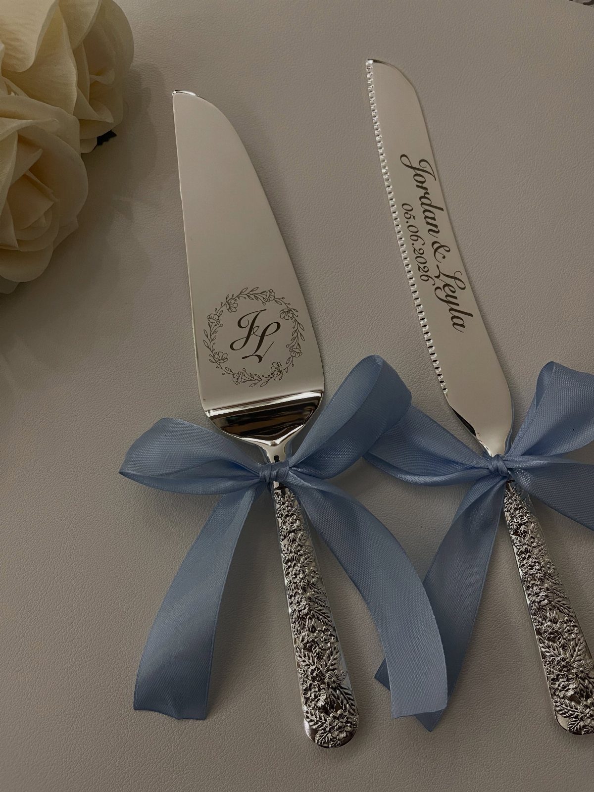 Engraved silver cake server and knife tied with light blue ribbons and white roses.