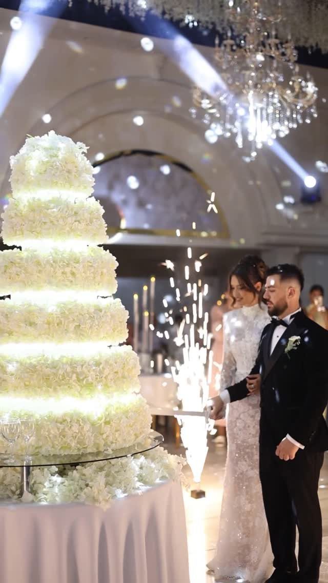 Bride and groom standing side by side next to a large white floral cake.