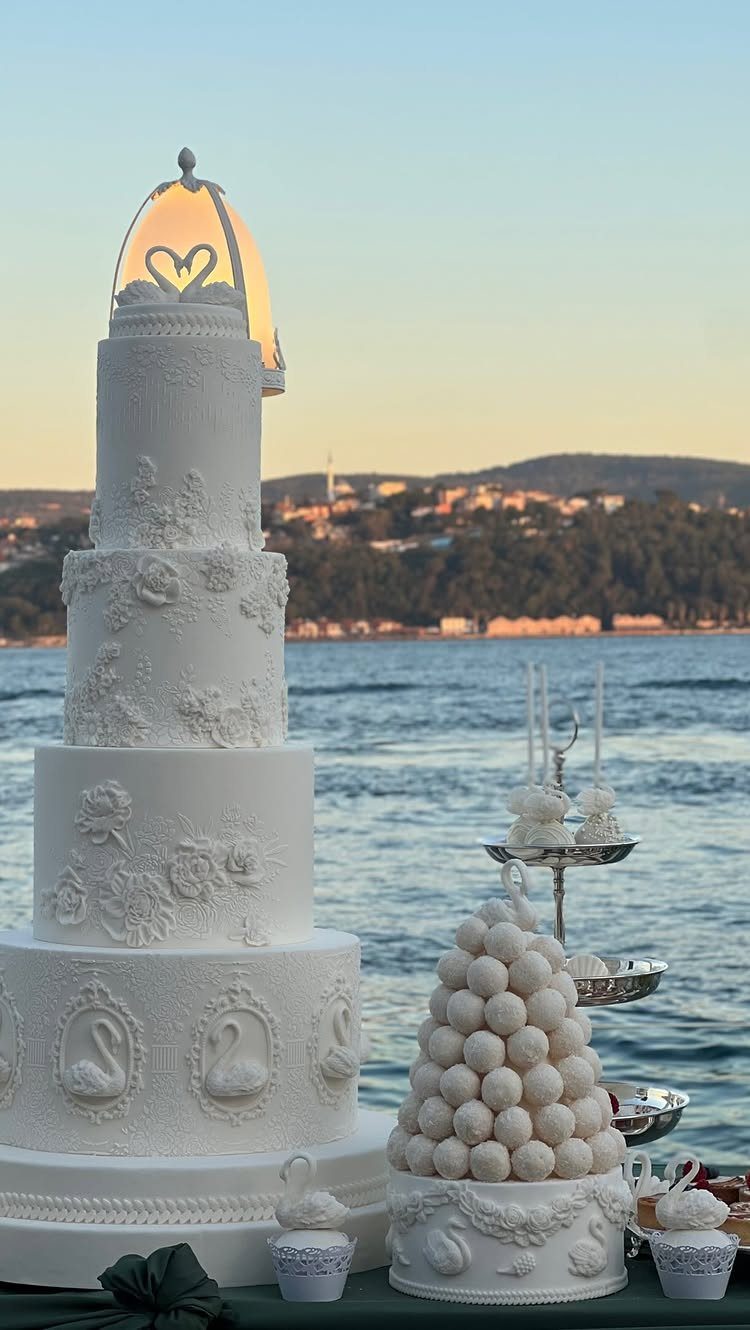 Five-tier white wedding cake with swan reliefs and a heart topper by the sea