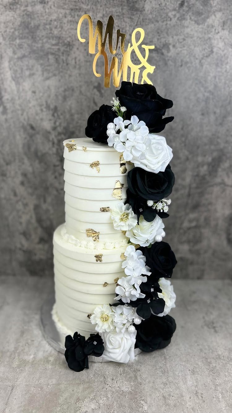 Two-tier white wedding cake decorated with cascading black and white roses and gold topper.