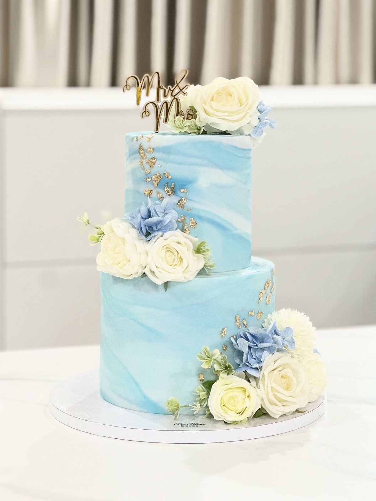Two-tier wedding cake with blue marbled icing, white roses, and gold topper.