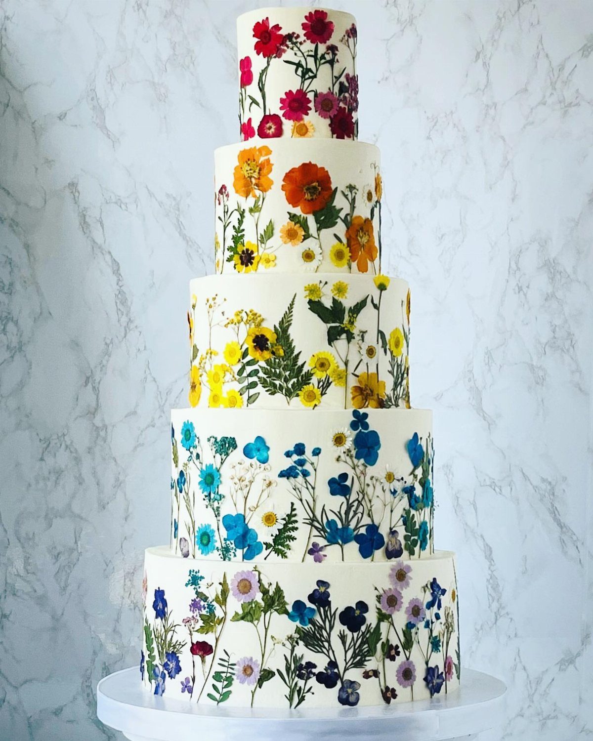 Five-tier white wedding cake decorated with colorful pressed flowers in a rainbow gradient.