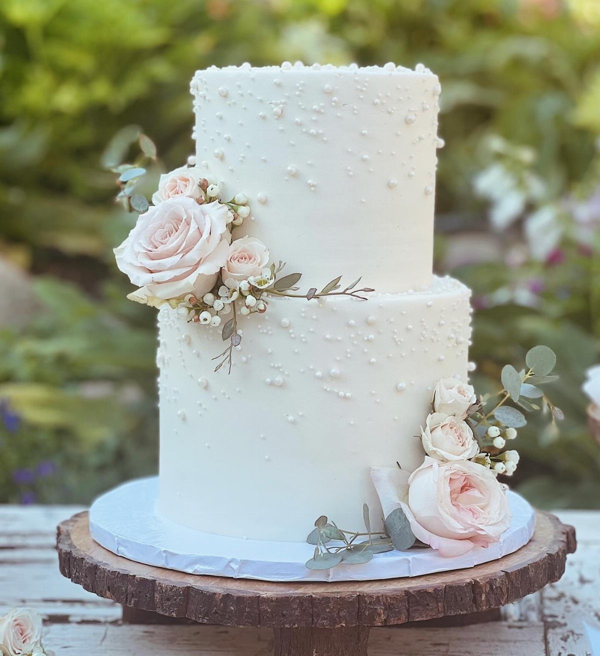 Two-tier white wedding cake with pearl decorations and pale pink roses on wood.