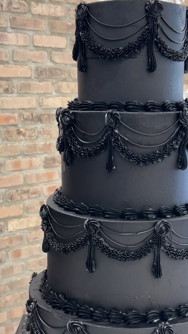 Multi-tiered black wedding cake with ornate frosting swags against a brick wall