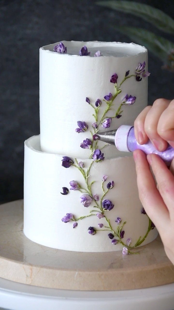 Two-tier white cake being decorated with purple buttercream flowers and green stems.