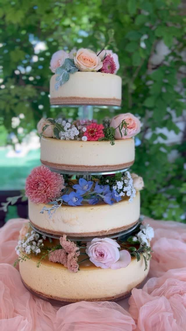 Four-tier cheesecake wedding cake decorated with fresh pink and blue flowers on pink fabric