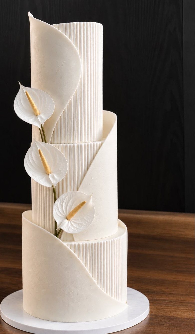 Three-tiered white wedding cake with ribbed textures, curved panels, and calla lily accents.