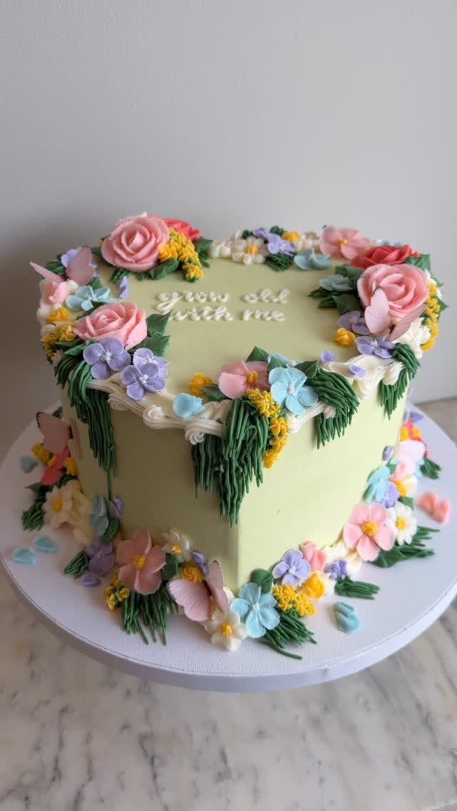Heart-shaped yellow cake decorated with colorful buttercream flowers and green leaves