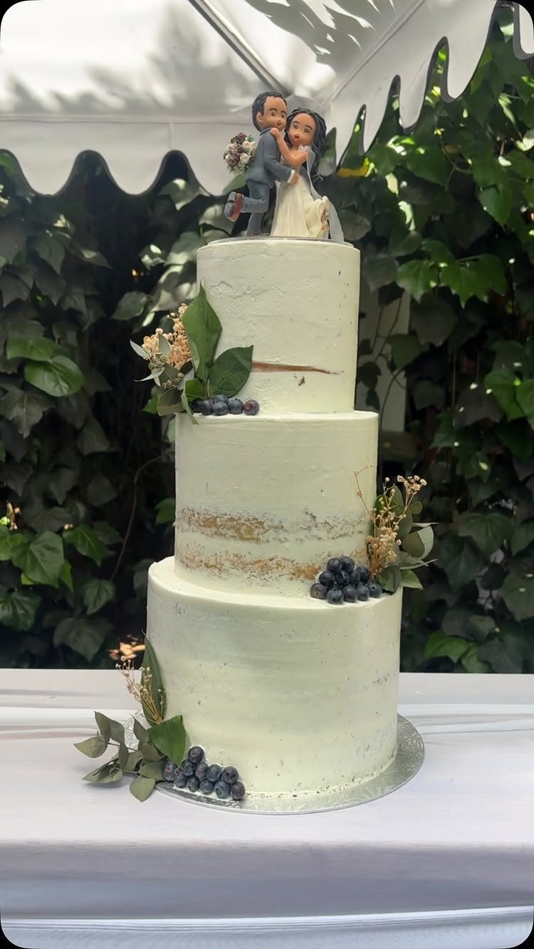 Three-tiered semi-naked wedding cake decorated with blueberries, greenery, and a couple topper.