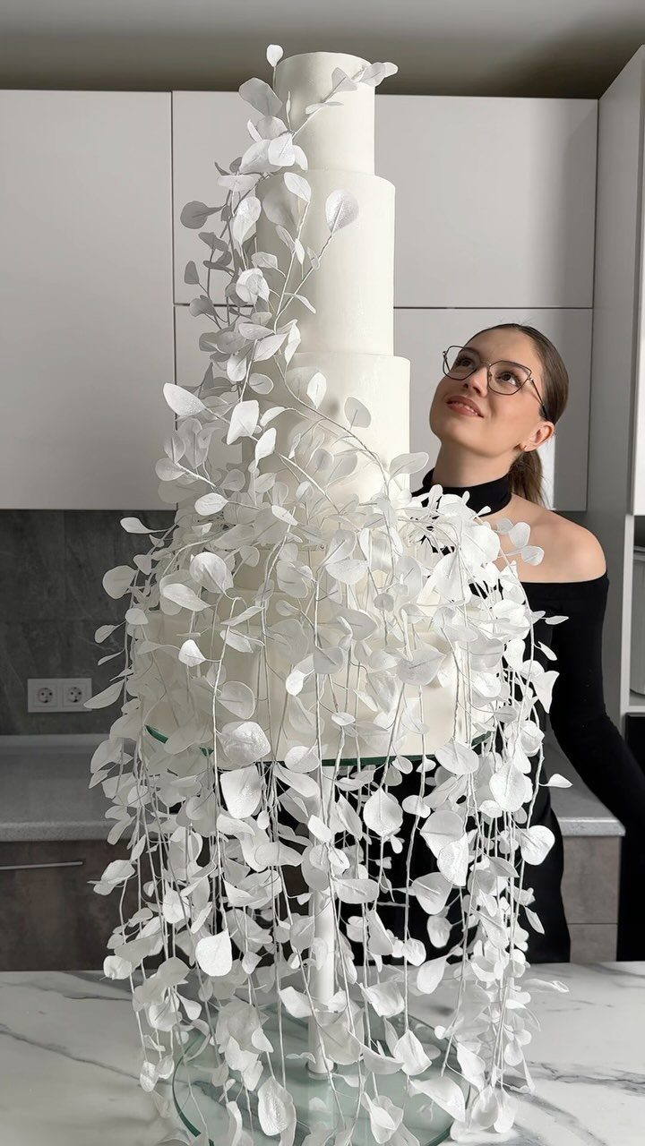Tall white tiered cake with cascading sugar leaves and a woman looking on.