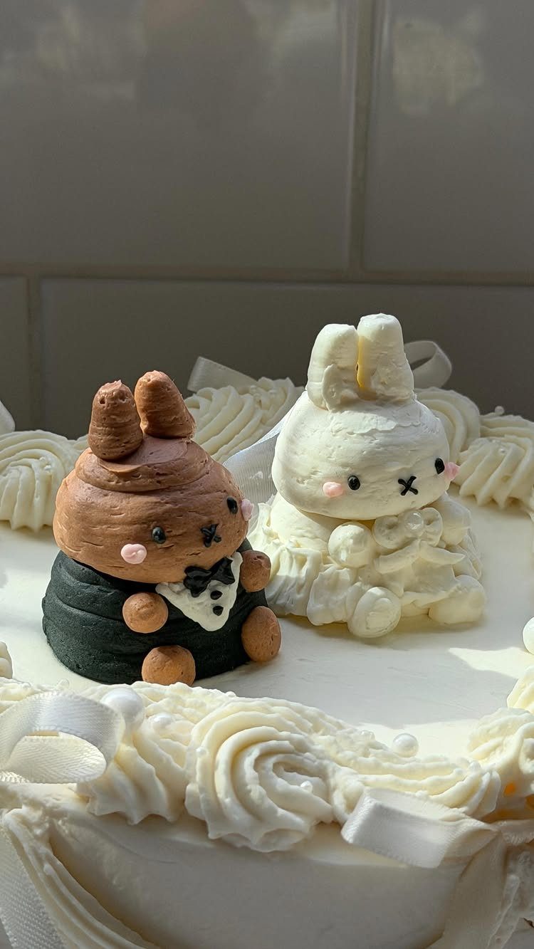 Two cute rabbit figurines in wedding attire sitting on a white frosted cake.