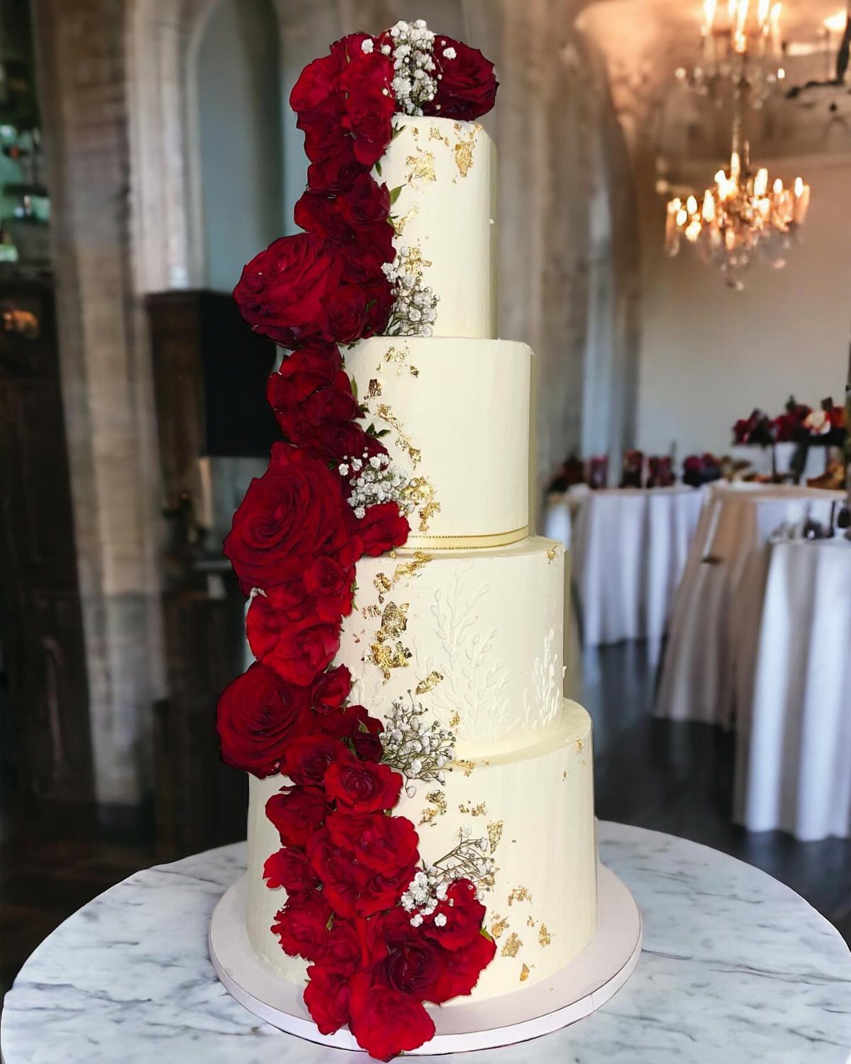 Four-tier white wedding cake with cascading red roses and gold leaf accents.