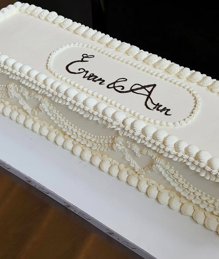 Rectangular white wedding cake with ornate frosting and names Evan and Ann written.