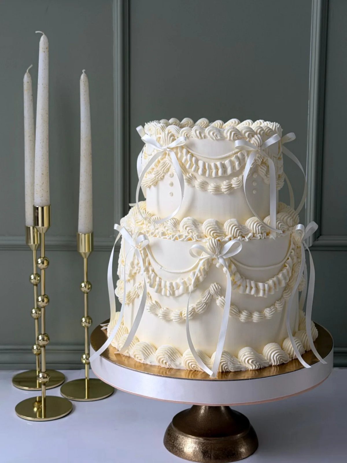 Two-tier white wedding cake with ornate piping and ribbons on a gold stand.