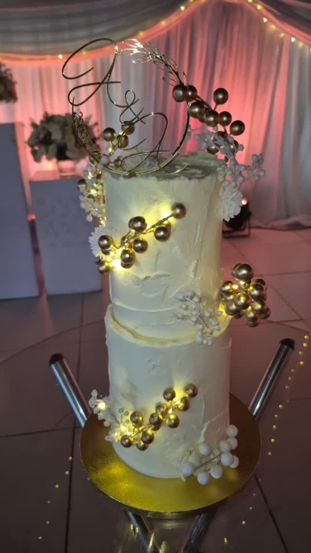 Two-tier white wedding cake decorated with gold spheres and glowing fairy lights.