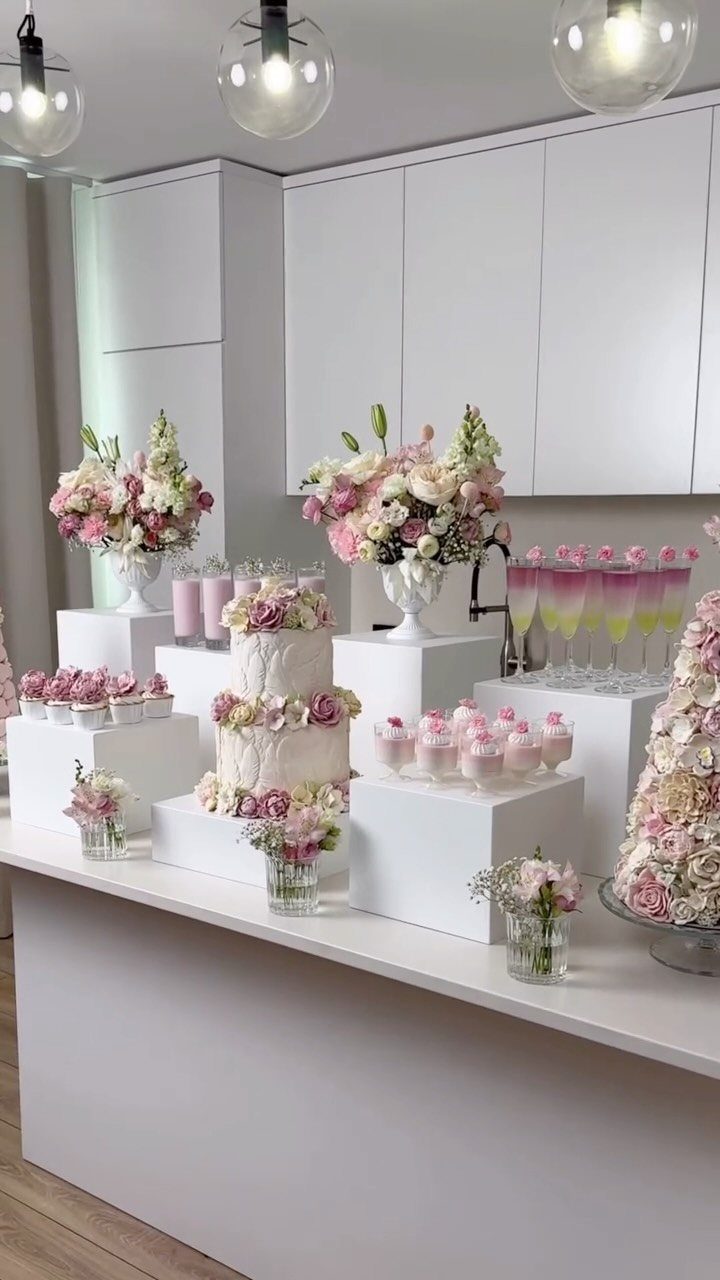 Wedding cake and pink cupcakes on a white dessert table with floral arrangements.