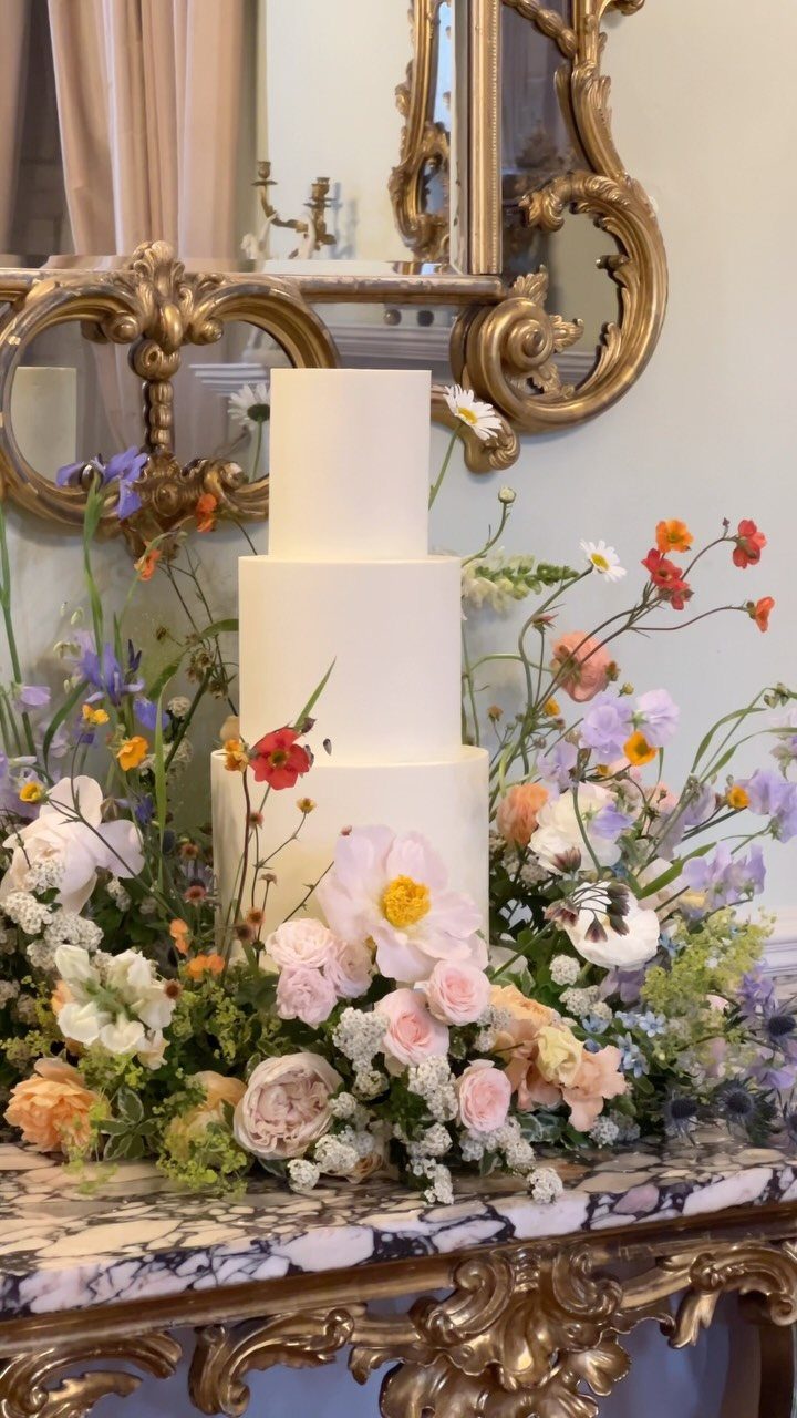 Three-tier white cake surrounded by colorful fresh flowers on a marble table.