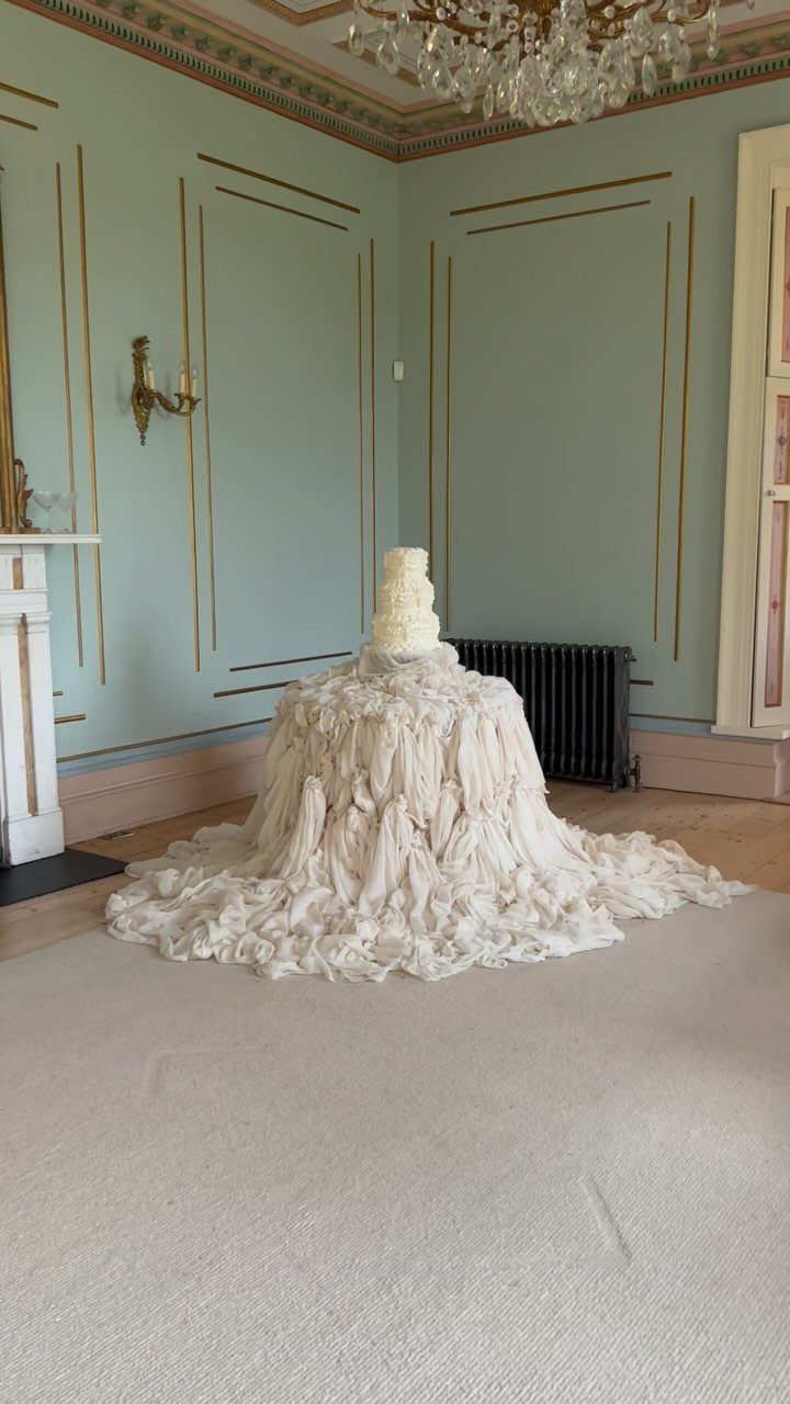 White wedding cake on a table covered in ruffled white fabric in a room.