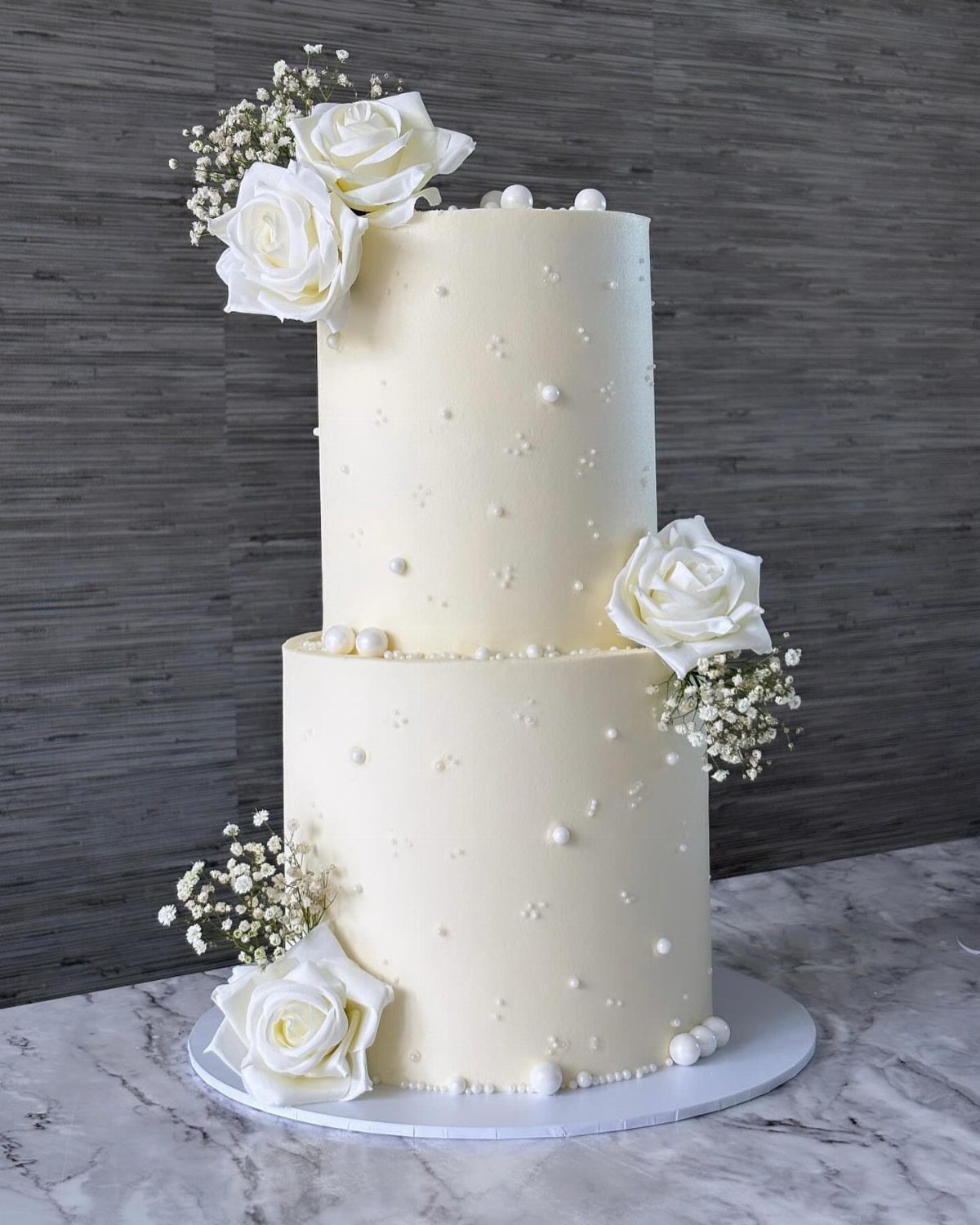 Two-tier white wedding cake with smooth frosting, white roses, and pearl decorations.