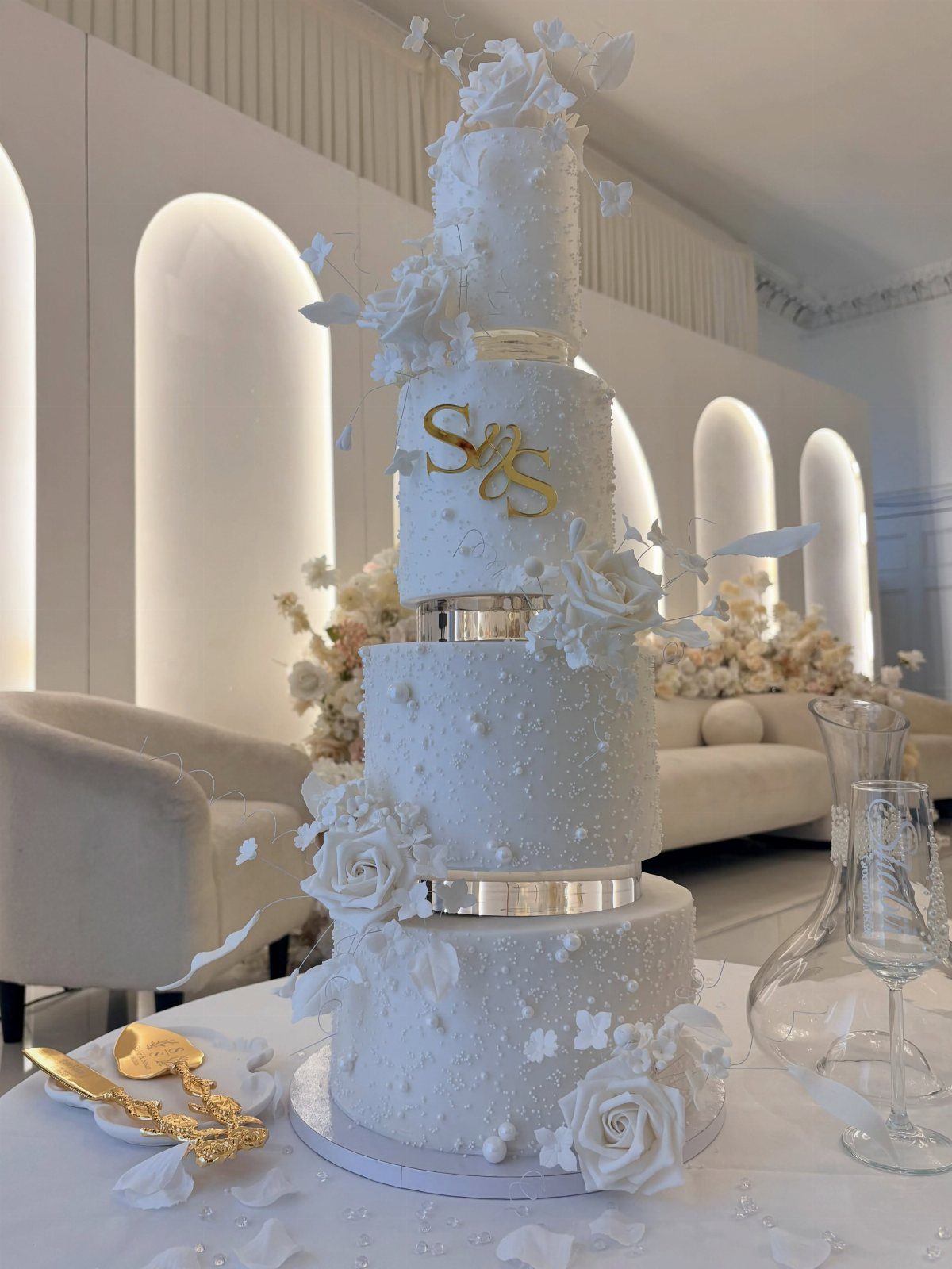 Four-tier white wedding cake with gold initials, silver bands, and white sugar roses.