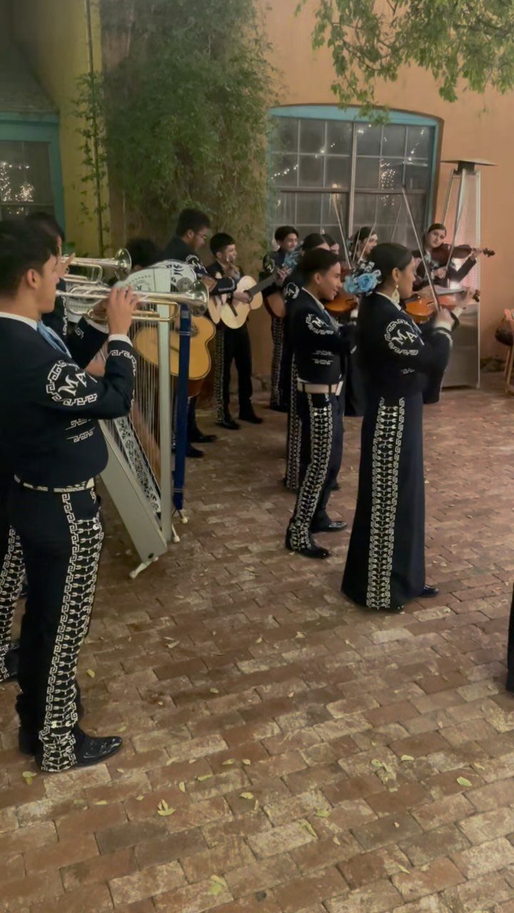Mariachi band in black charro suits playing instruments on a brick patio.