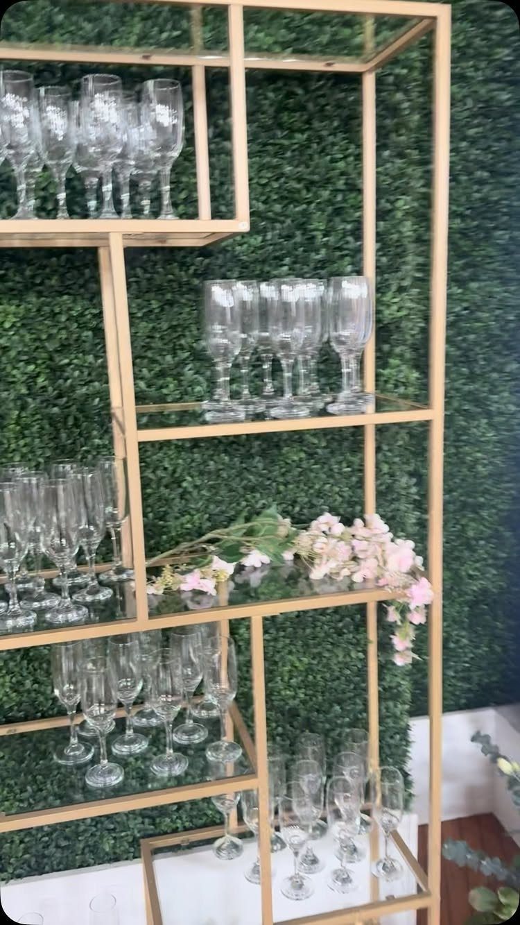 Gold geometric shelves with champagne flutes against a green hedge wall with flowers