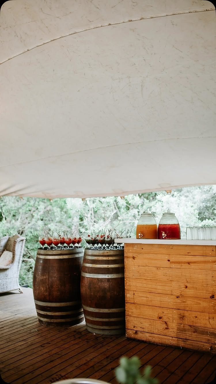 Wooden bar with drink dispensers and glasses on barrels under a white canopy