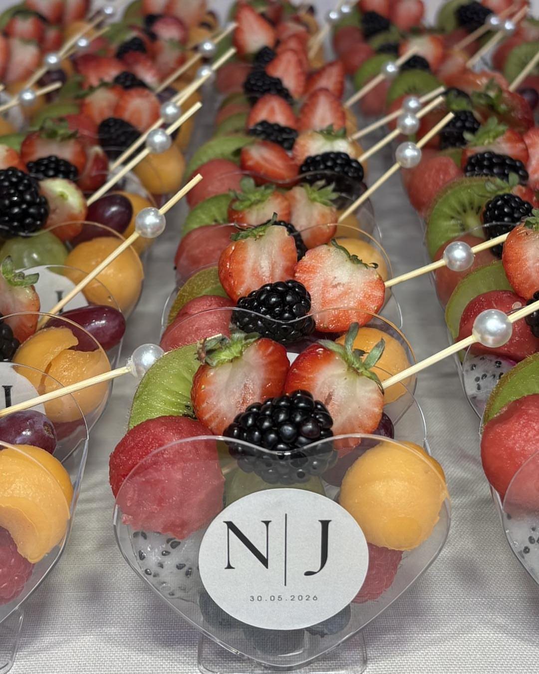 Fruit skewers with watermelon, cantaloupe, kiwi, strawberries, and blackberries in clear plastic cups.
