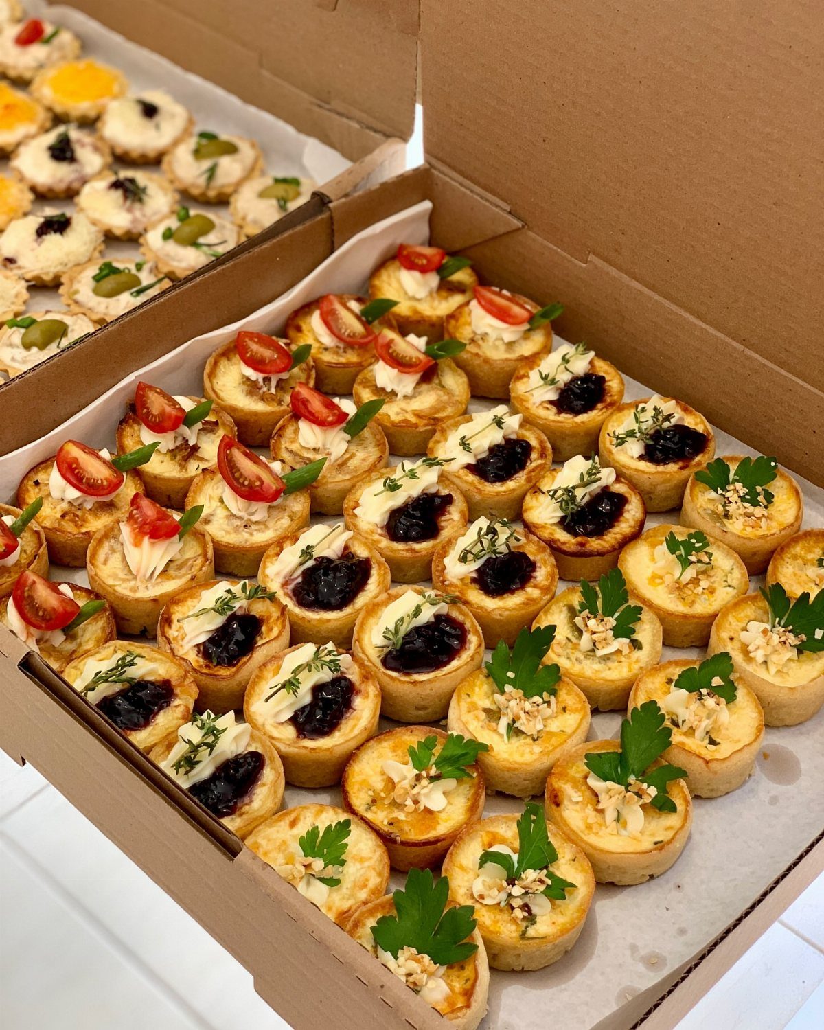 Assorted mini quiches with tomato, berry, and herb toppings in cardboard boxes.